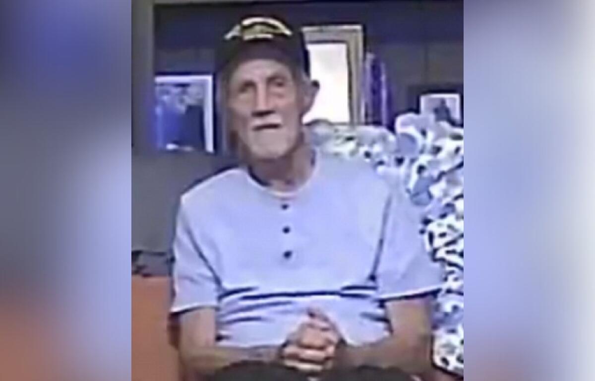 Elderly man wearing a light blue shirt and Navy veteran hat, seated indoors with hands folded.