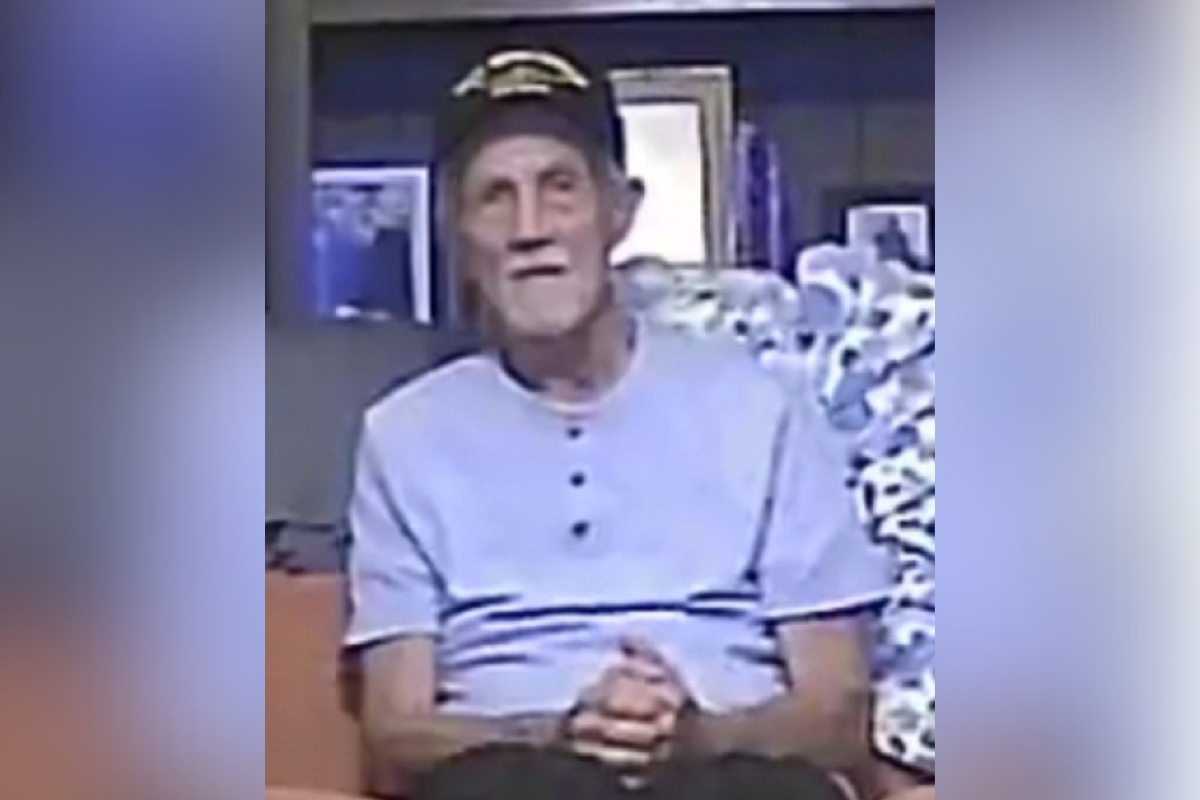Elderly man wearing a light blue shirt and Navy veteran hat, seated indoors with hands folded.