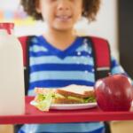 Child holding a red lunch tray with a sandwich, apple, and bottle of milk at a school meal program.