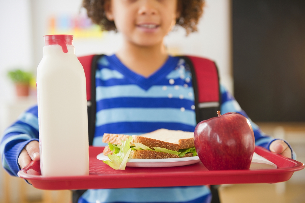 Child holding a red lunch tray with a sandwich, apple, and bottle of milk at a school meal program.