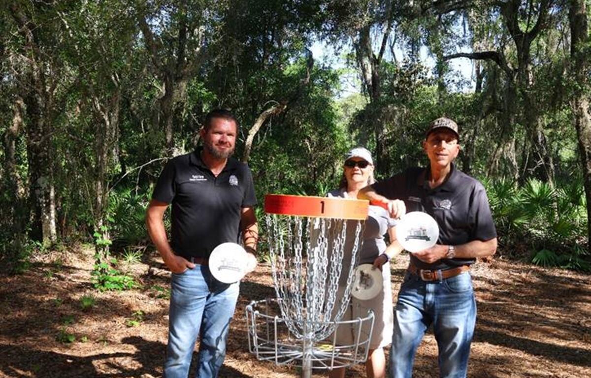 Sumter County Commissioners Coon, Butterfield, and Bilardello stand beside a disc golf basket at Lake Okahumpka Park, each holding a disc during the course’s ribbon-cutting ceremony.