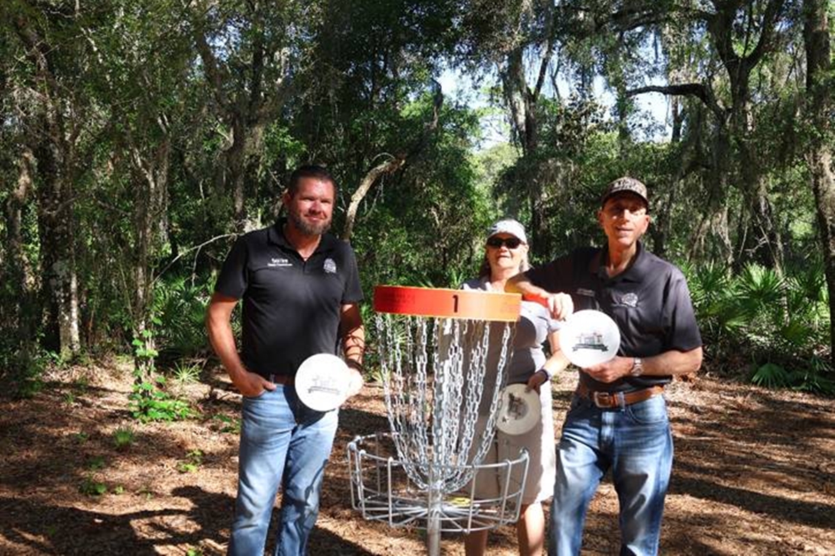 Sumter County Commissioners Coon, Butterfield, and Bilardello stand beside a disc golf basket at Lake Okahumpka Park, each holding a disc during the course’s ribbon-cutting ceremony.