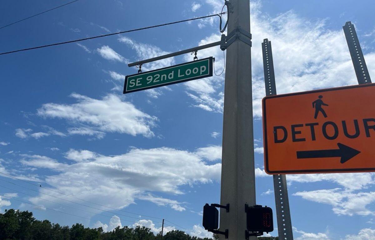 A street sign attached to a concrete pole, with several other attachments, with blue skies, with white puffy clouds to the right, and a smaller pole with an orange sign to the left.