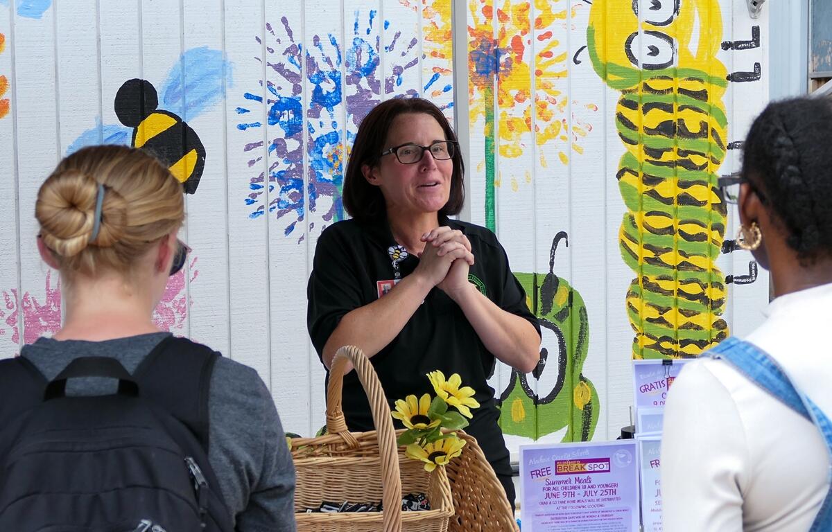 Jamie Lovett talks to community members at a Summer Meals kickoff event, standing in front of a brightly painted wall with flowers and insects.