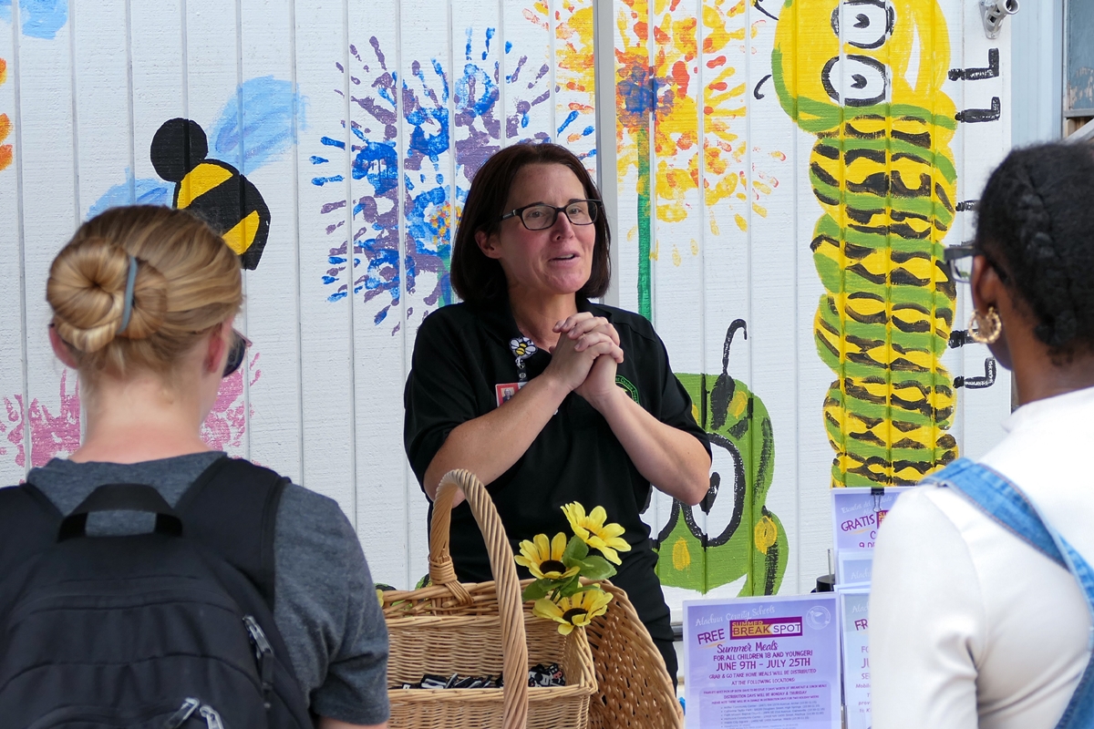 Jamie Lovett talks to community members at a Summer Meals kickoff event, standing in front of a brightly painted wall with flowers and insects.