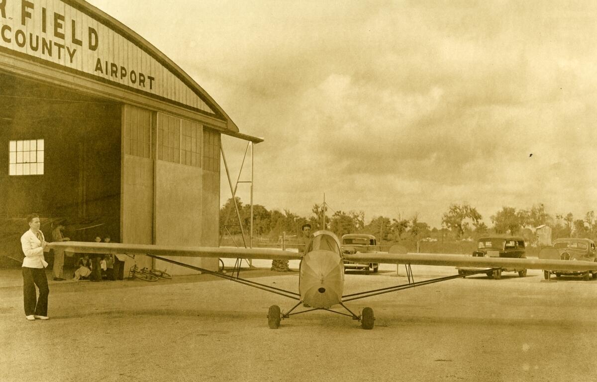 Sepia-toned photograph of a small glider in front of the Taylor Field hangar at Ocala-Marion County Airport. The sleek, narrow aircraft is parked on the tarmac, with a man standing at the end of one wing. The hangar doors are open, with several people sitting just inside. Vintage automobiles are parked in the background under a cloudy sky, giving the scene a mid-20th-century atmosphere.
