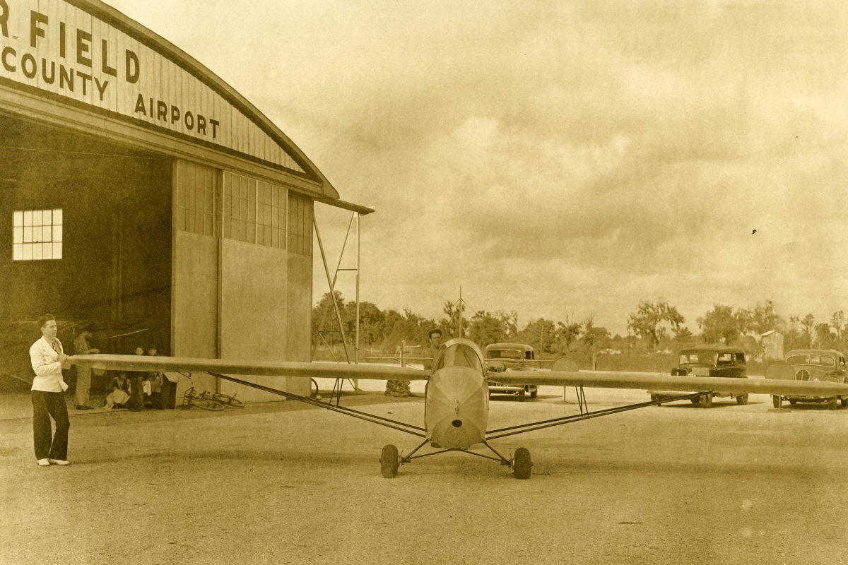 Sepia-toned photograph of a small glider in front of the Taylor Field hangar at Ocala-Marion County Airport. The sleek, narrow aircraft is parked on the tarmac, with a man standing at the end of one wing. The hangar doors are open, with several people sitting just inside. Vintage automobiles are parked in the background under a cloudy sky, giving the scene a mid-20th-century atmosphere.
