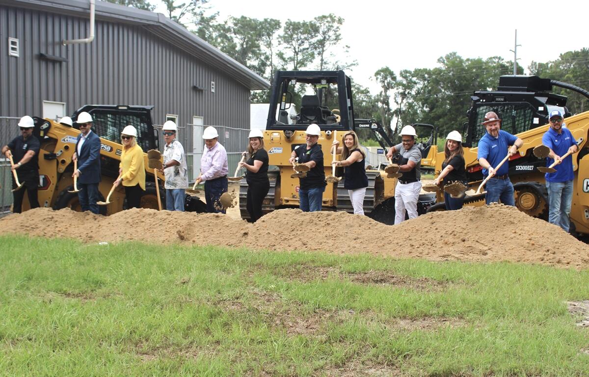 Group of people in hard hats using shovels to break ground in front of construction equipment at Transitions Life Center expansion site.