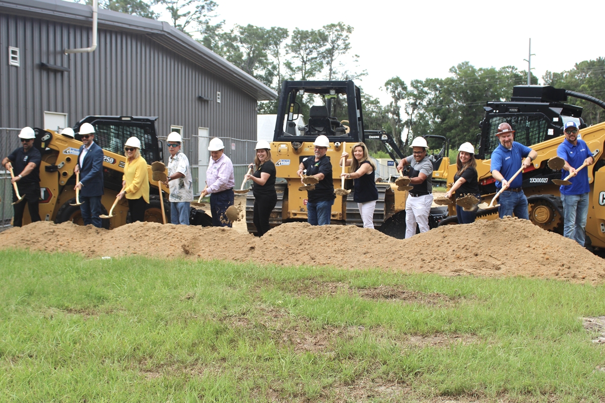 Group of people in hard hats using shovels to break ground in front of construction equipment at Transitions Life Center expansion site.
