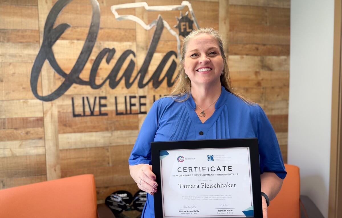 Tamara Fleischhaker smiling and holding a framed workforce development certificate in front of a wood-paneled wall with the Ocala logo.