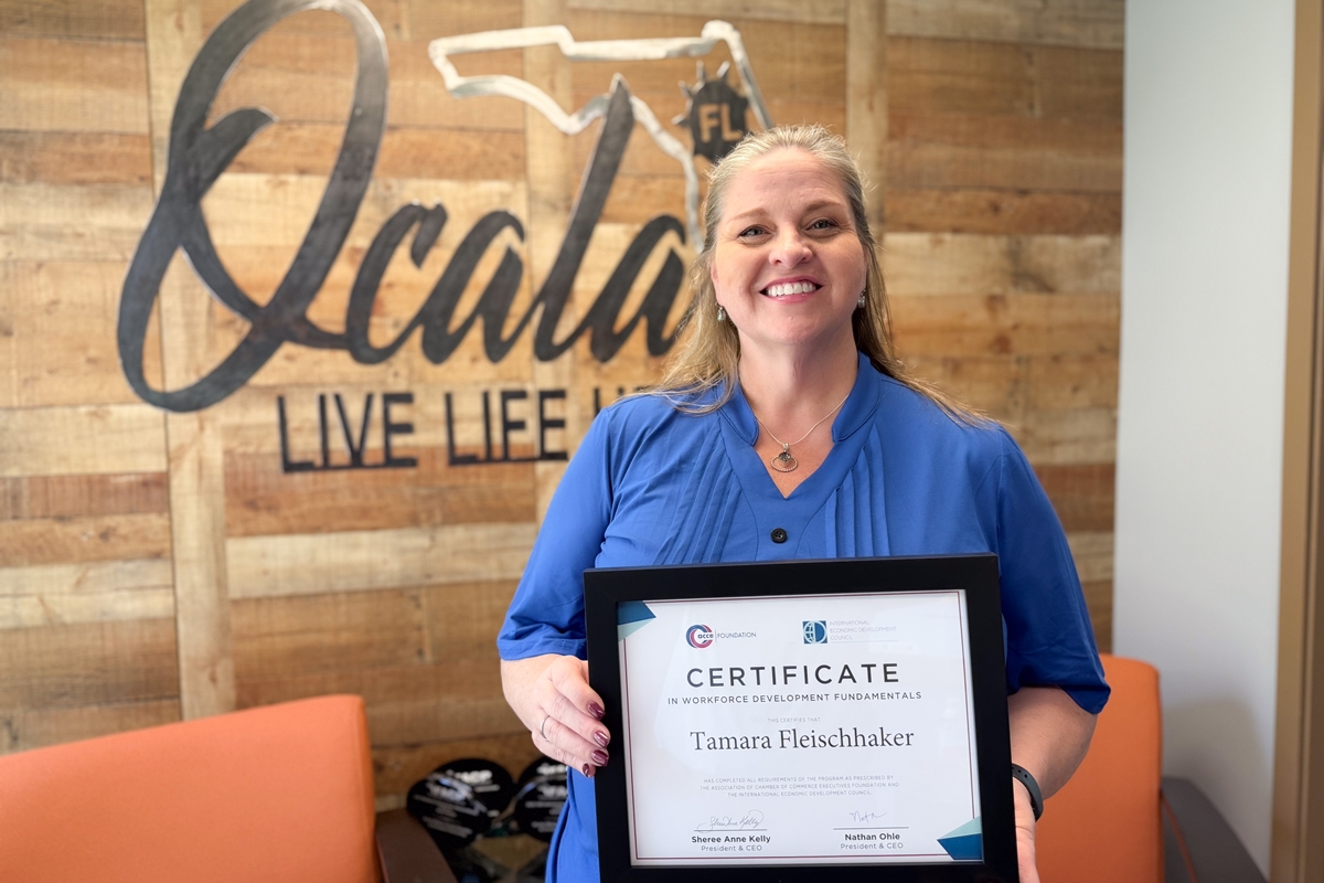 Tamara Fleischhaker smiling and holding a framed workforce development certificate in front of a wood-paneled wall with the Ocala logo.