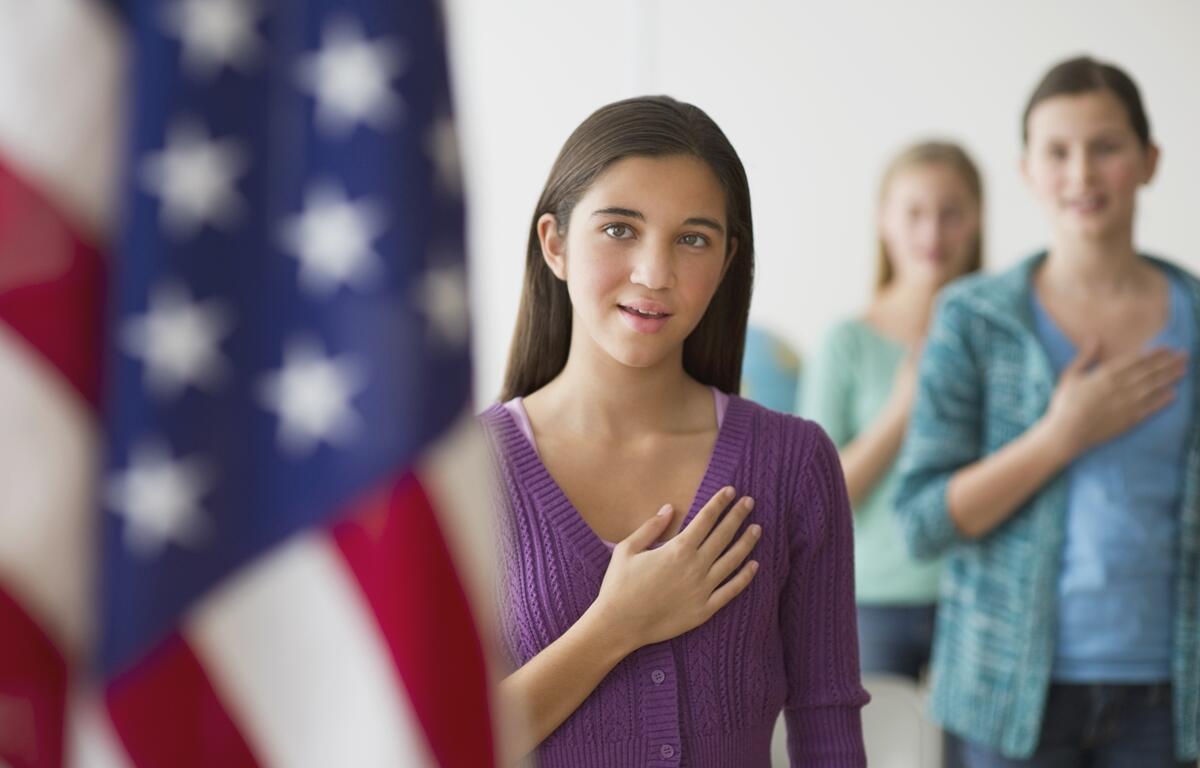 Three high school girls with hands over their hearts reciting the Pledge of Allegiance in front of an American flag.