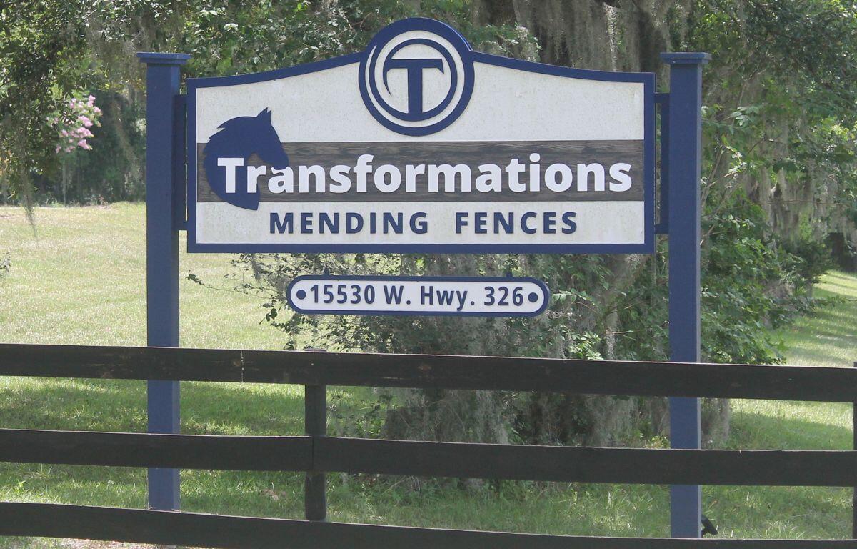 A white sign, with blue lettering, with a three-board fence in the foreground, and grass and live oak trees with Spanish moss in the background.