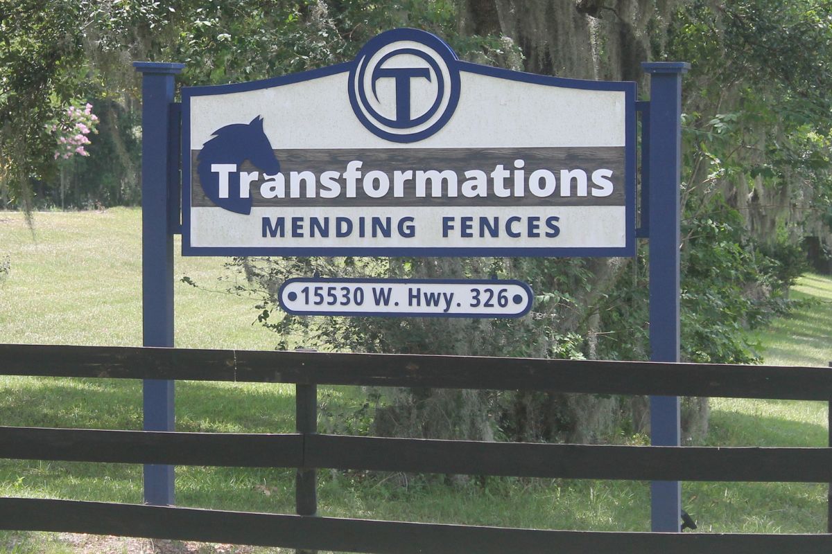 A white sign, with blue lettering, with a three-board fence in the foreground, and grass and live oak trees with Spanish moss in the background.