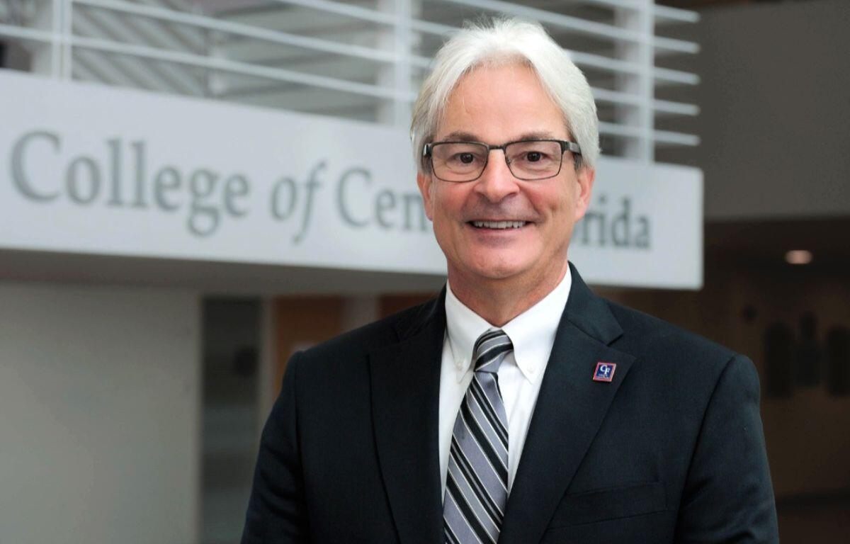 A professionally dressed man with white hair and glasses smiling in front of a sign that reads 'College of Central Florida.' He is wearing a dark suit, striped tie, and a lapel pin.