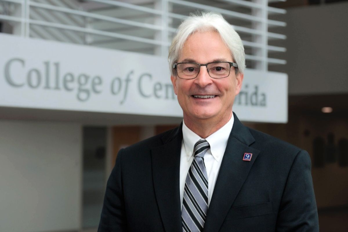 A professionally dressed man with white hair and glasses smiling in front of a sign that reads 'College of Central Florida.' He is wearing a dark suit, striped tie, and a lapel pin.