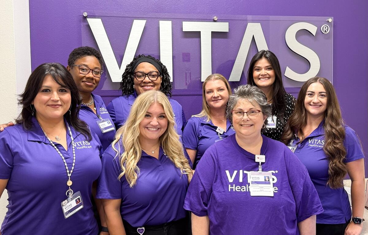 Eight smiling VITAS Healthcare staff members wearing purple uniforms stand in front of a VITAS sign inside the new Ocala office.