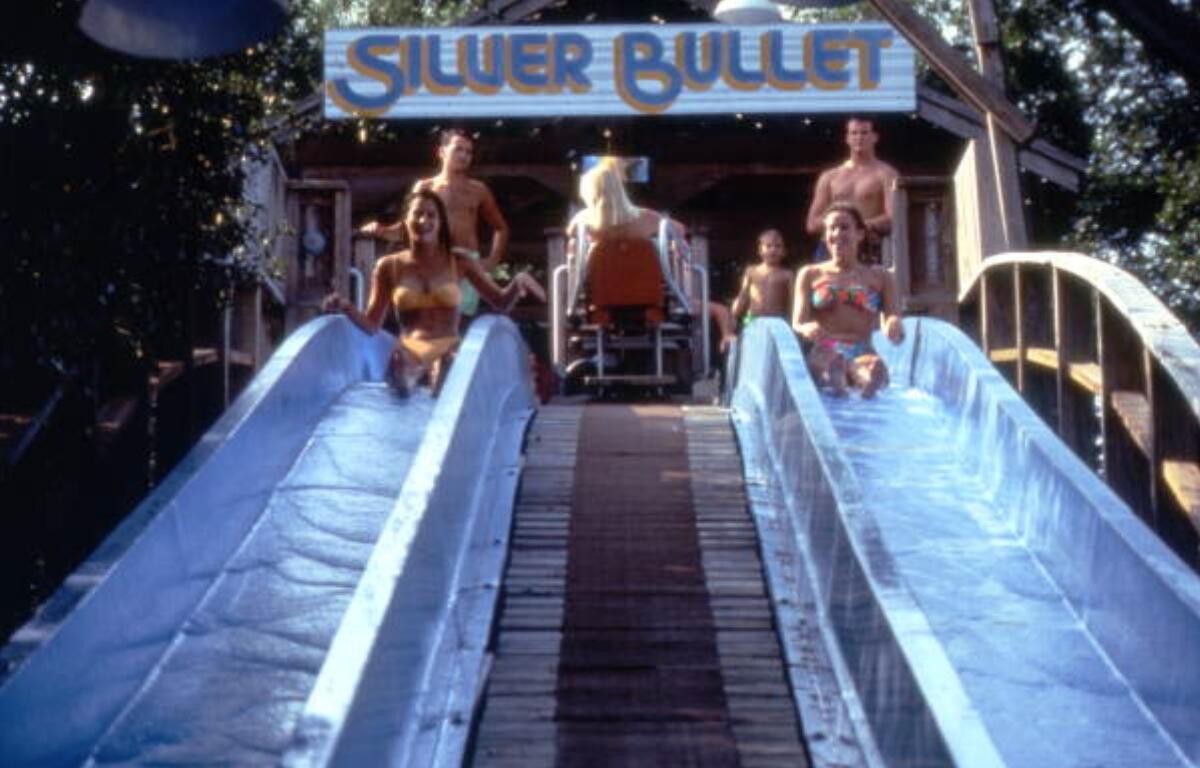 A group of people prepare to slide down a pair of water slides labeled "Silver Bullet" at a water park. Two women in swimsuits are seated at the top of the slides, smiling. Behind them, other individuals, including a person in an orange and white wheelchair, wait their turn. The scene is surrounded by trees, and the slide structure is wooden with a sign overhead.