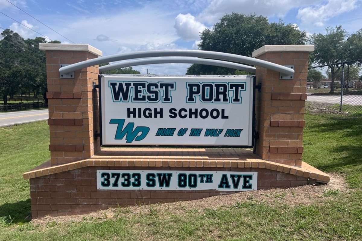 A white sign with lettering between two brown colored standards, with a small rectangular sign with letters and numbers, with a blue sky with clouds and a green tree in the background. 