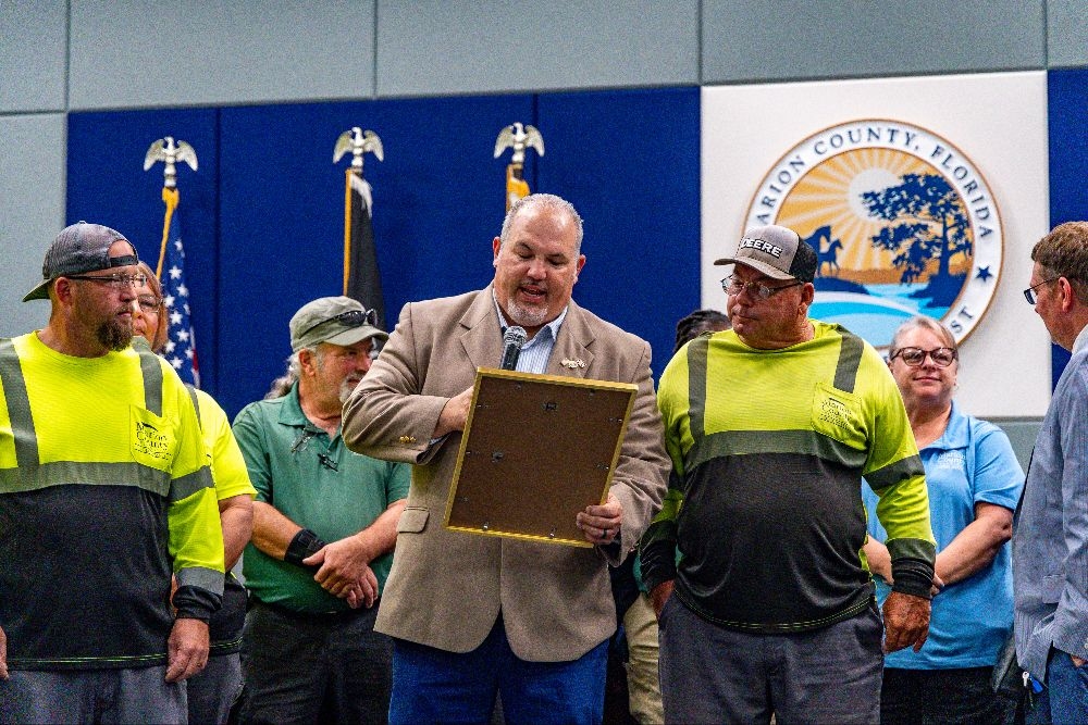 Carl Zalak III reads a framed proclamation surrounded by waste management workers and county staff at a Marion County Commission meeting.
