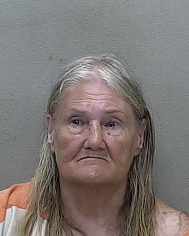 mugshot of an older woman with long, light gray hair, wearing a striped orange and white garment, against a plain background.