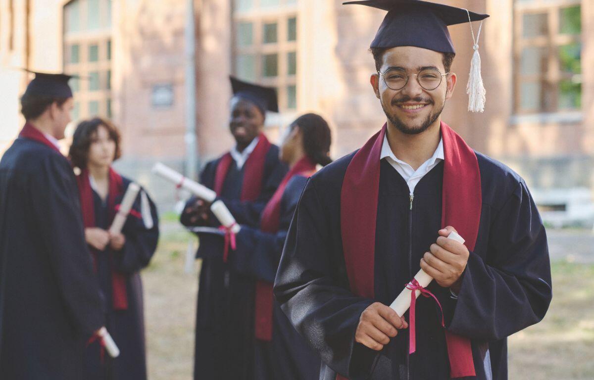 A group of graduates in caps and gowns, holding diplomas, celebrates their achievement outdoors, with one smiling graduate prominently in the foreground.
