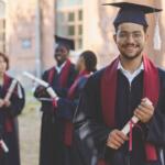 A group of graduates in caps and gowns, holding diplomas, celebrates their achievement outdoors, with one smiling graduate prominently in the foreground.