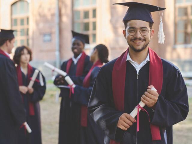 A group of graduates in caps and gowns, holding diplomas, celebrates their achievement outdoors, with one smiling graduate prominently in the foreground.