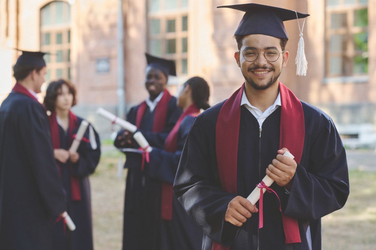 A group of graduates in caps and gowns, holding diplomas, celebrates their achievement outdoors, with one smiling graduate prominently in the foreground.