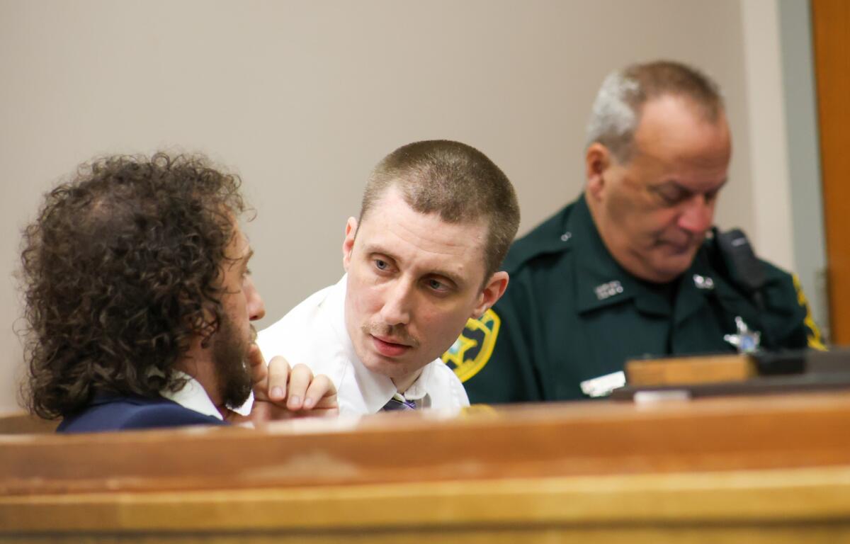 A man in a white shirt looks over to his attorney in a blue suit, while a police officer sits in the background