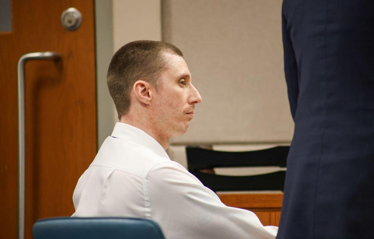 A man in a white dress shirt is seated in a courtroom, appearing serious and attentive as he looks forward.