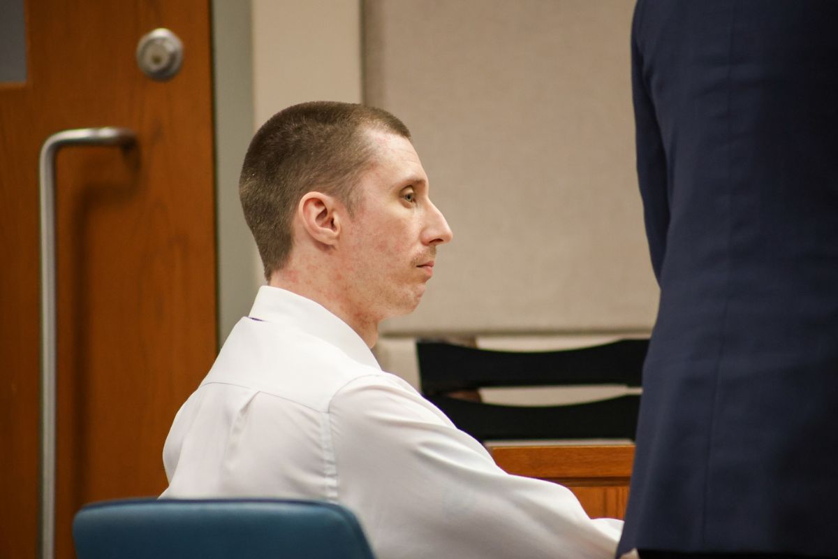 A man in a white dress shirt is seated in a courtroom, appearing serious and attentive as he looks forward.