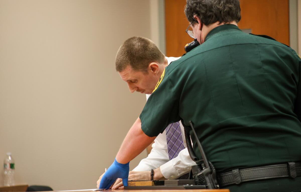 A man in a white shirt looks down at a table while a police officer in a green uniform instructs him on what to do