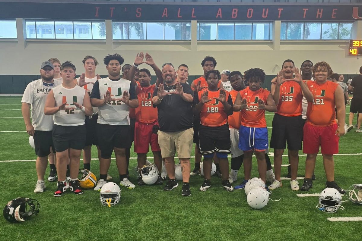 A group of young athletes pose for a photo inside an indoor football facility, all wearing orange "U" shirts and helmets, indicating participation in a linemen training camp. The atmosphere is upbeat and focused, with players in the background prepping for drills.