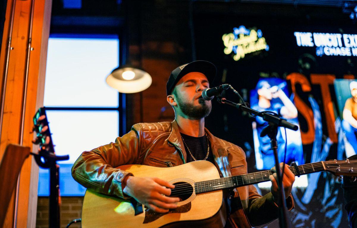 Musician performs with guitar in hand and in front of a microphone in a darker venue with lights in the background