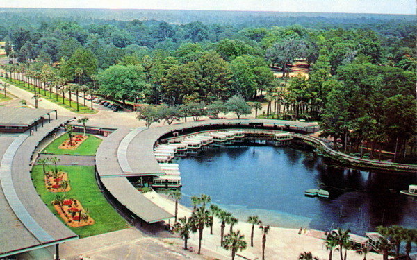Aerial view of the boat docks at Florida’s Silver Springs, featuring a circular lagoon lined with covered walkways and glass-bottom boats. The surrounding area is filled with lush green trees and landscaped flower beds, with palm trees lining the walkways. The background shows a dense forest stretching to the horizon.