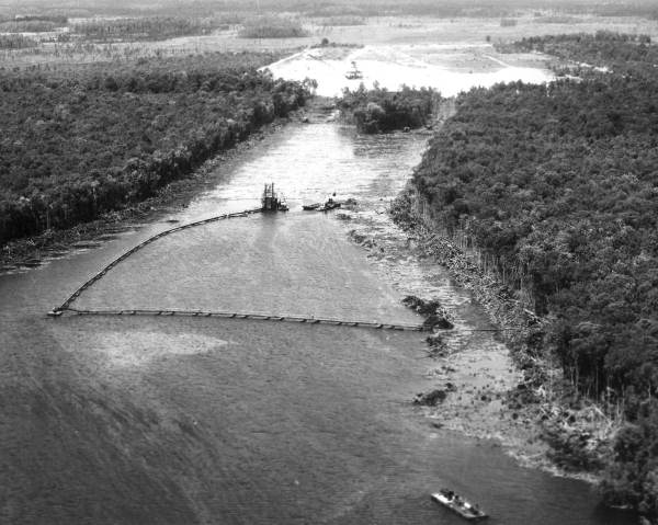 A black-and-white aerial view of construction. An excavated channel filled with water is carved through a forested area as boats carry building materials.