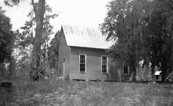 A black-and-white photo of a small wooden church or schoolhouse with a steep metal roof, raised slightly above the ground. The simple rectangular building has tall, narrow windows along its side and is surrounded by tall grass, trees, and Spanish moss. The setting appears rural and quiet, with no people visible.