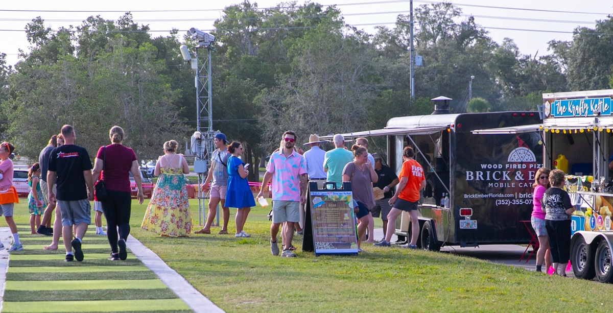 Parents and children gather at food trucks serving kettle corn and wood-fired pizza.