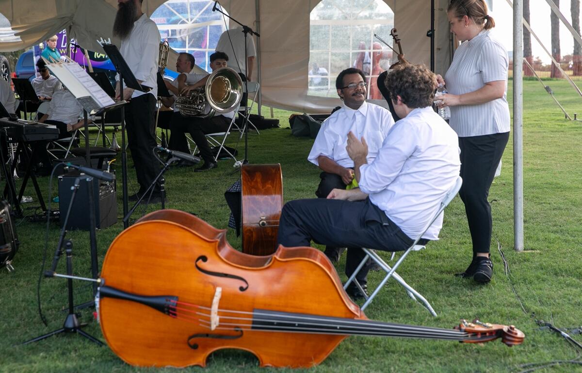 Musicians in white shirts relax and chat behind the performance tent with instruments nearby during Symphony Under the Stars.