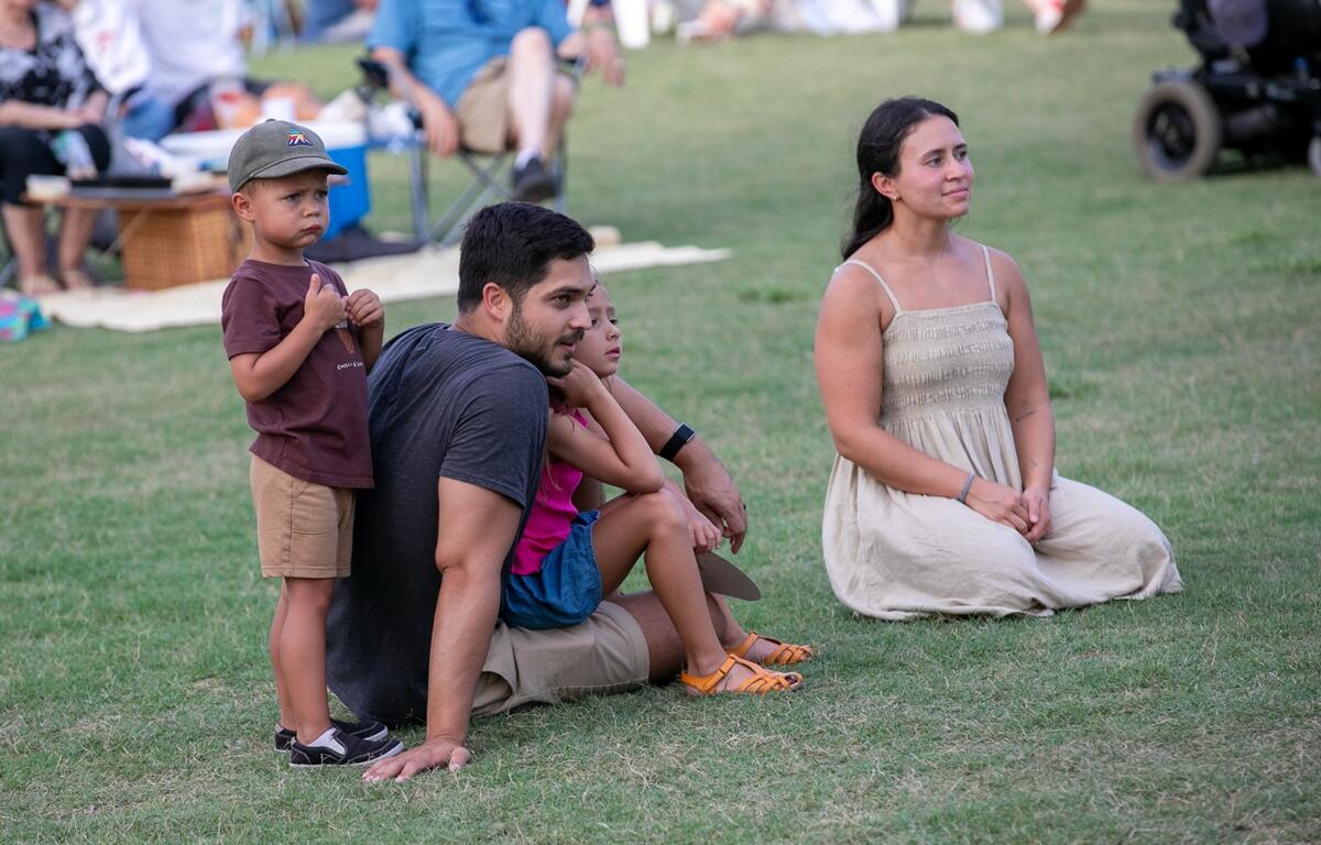 Two adults and three children sit on the grass, watching the stage with interest.