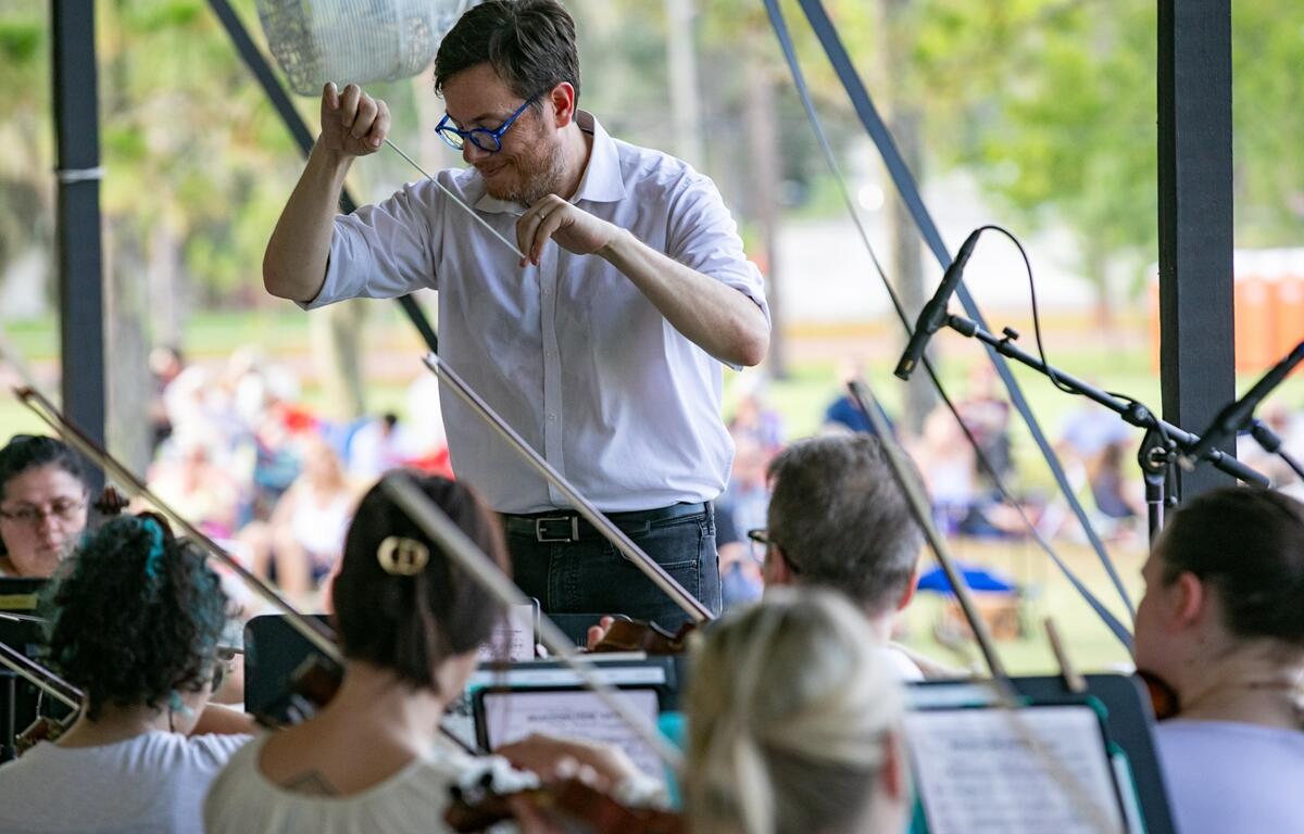 Matt Wardell conducts musicians in an outdoor concert with trees and tents in the background.