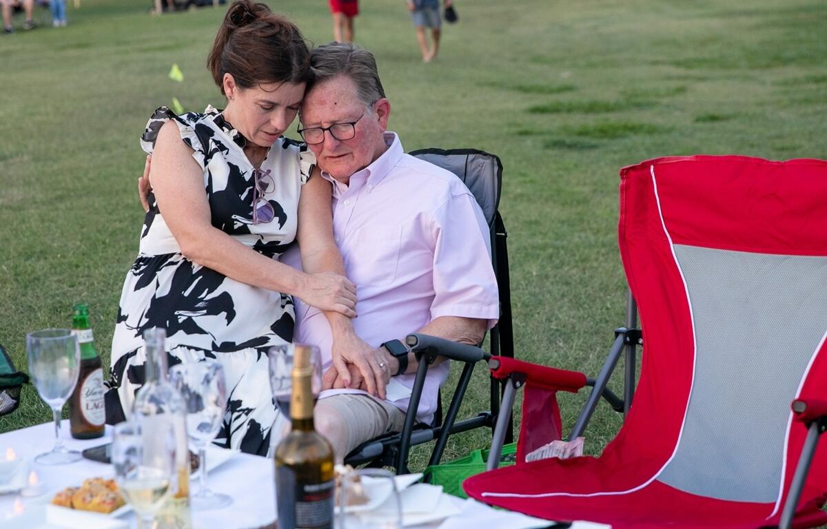 A woman embraces a man at a table set with wine and snacks on the lawn during the evening performance.
