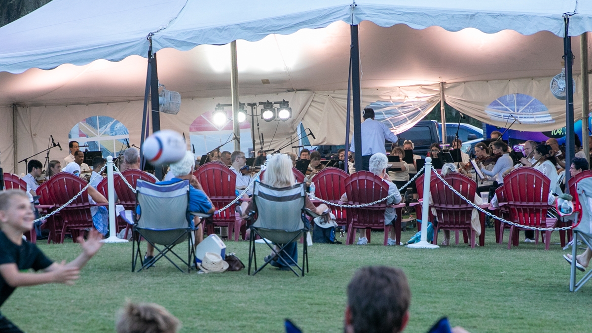 Rows of red chairs face the stage under a white tent as the orchestra performs at sunset.
