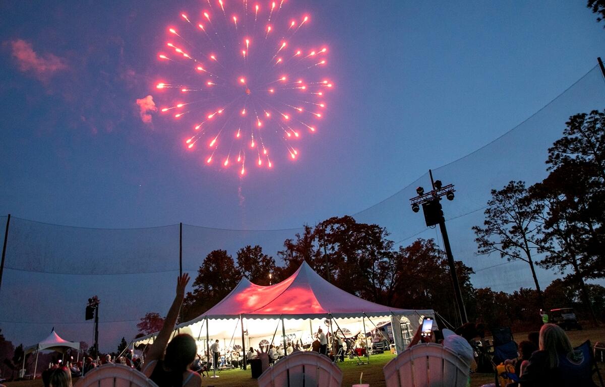 Audience members in lawn chairs look up at a red firework bursting above the concert tent at dusk.
