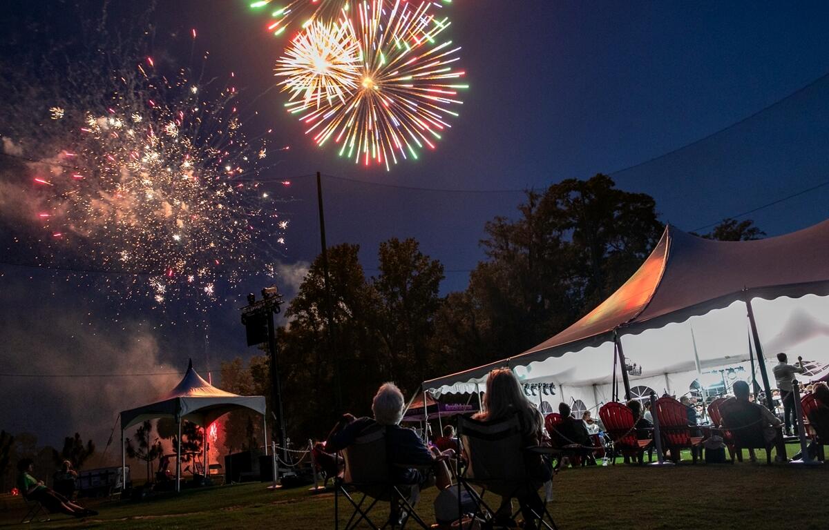 Multicolored fireworks illuminate the sky while guests in chairs enjoy the orchestra's performance.