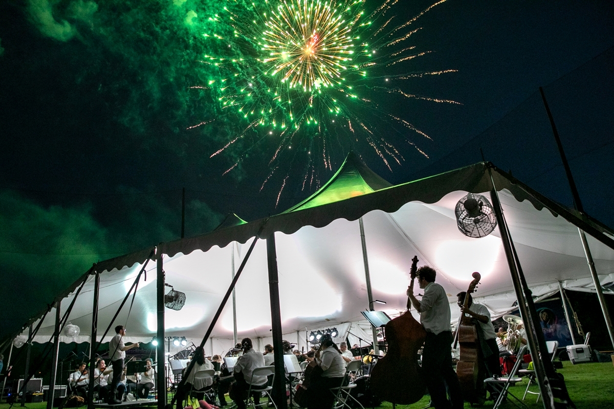 Green and gold fireworks shine above the white concert tent as the orchestra performs underneath.