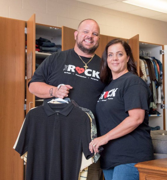 A man and woman stand together in what appears to be a clothing donation center, holding up several shirts on hangers. Both are smiling and wearing matching shirts with the logo “THE ROCK” and the phrase “Refuge · Foundation · Purpose,” indicating their involvement in a charitable or community support organization.
