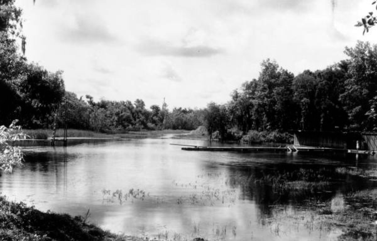 Black and white photo of a calm river bordered by dense trees on both sides. A wooden dock or platform stretches into the water on the right, with a small figure standing nearby. Reflections of clouds and vegetation are visible in the water, suggesting a peaceful, rural setting.