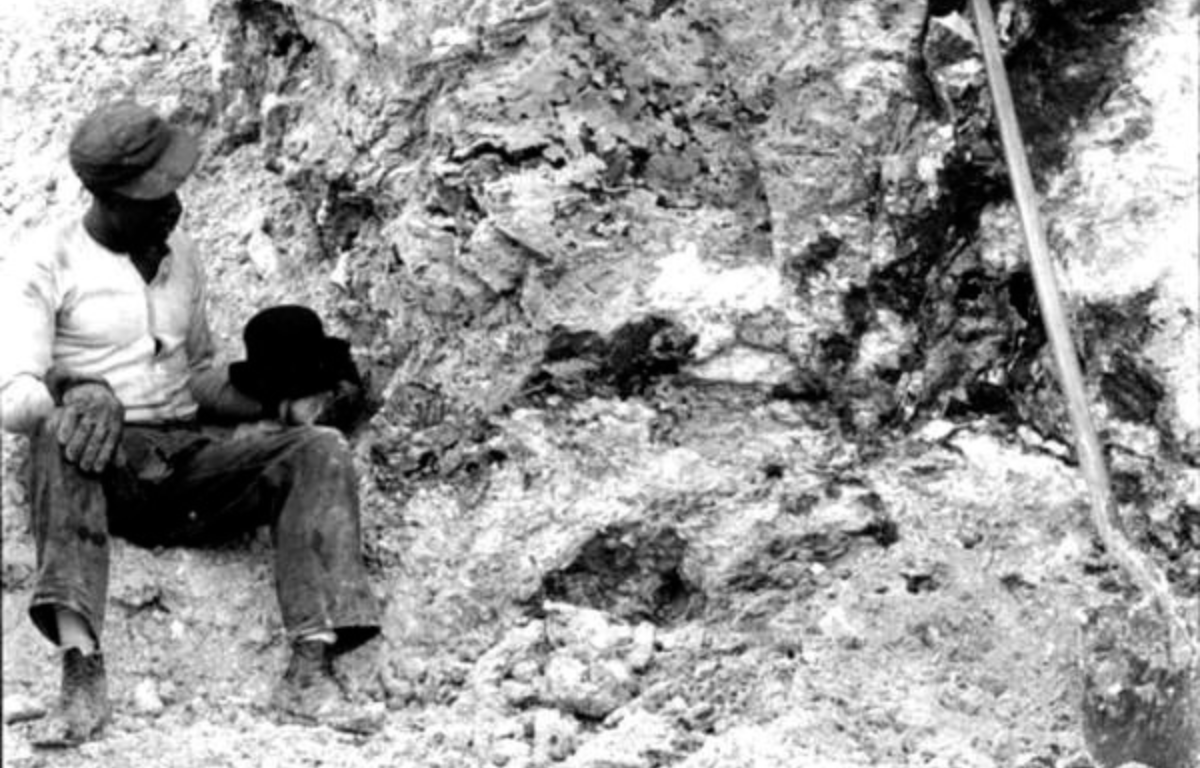 Black and white photo of a man sitting against a rocky embankment, likely in a phosphate mine. He wears work clothes, boots, and a cap, and holds a hat in one hand. A shovel is propped up beside him, and the textured rock wall behind him indicates active or recent excavation.